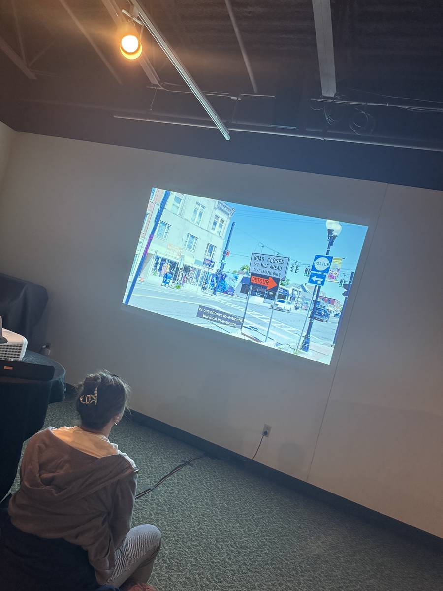 A visitor sitting on the floor of a dark room watching a projected film of a Johnson City street with road-closed and detour signs, with a subtitle about out-of-town investment.